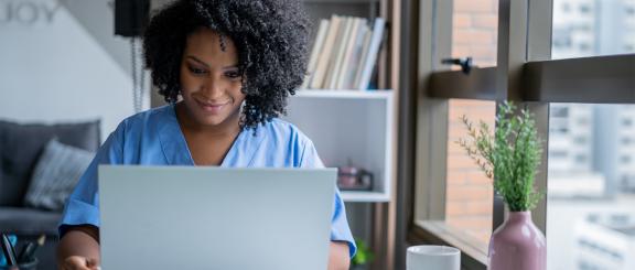 Woman in nurse scrubs looking at computer in her home.