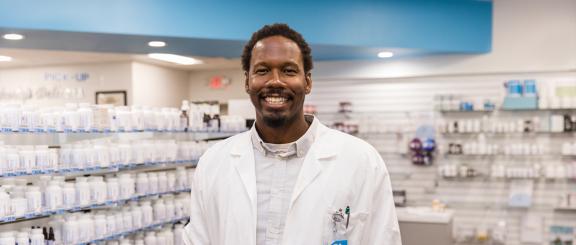Man in white lab coat smiling in front of shelves of white medicine bottles.
