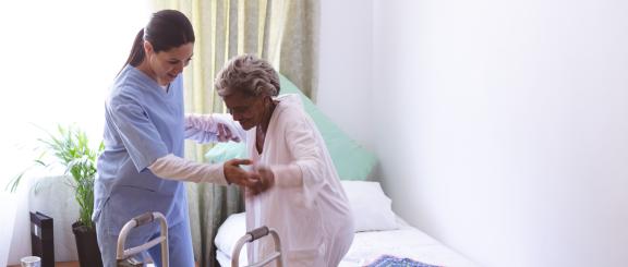 A younger palliative care worker wearing blue scrubs helps an elderly patient stand up from her bed to get to her walker. Both people are smiling as they interact and the room is brightly lit.