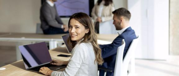 Smiling woman sits at long conference room desk with laptop. Other professionals in the background in front of a board with a line graph.